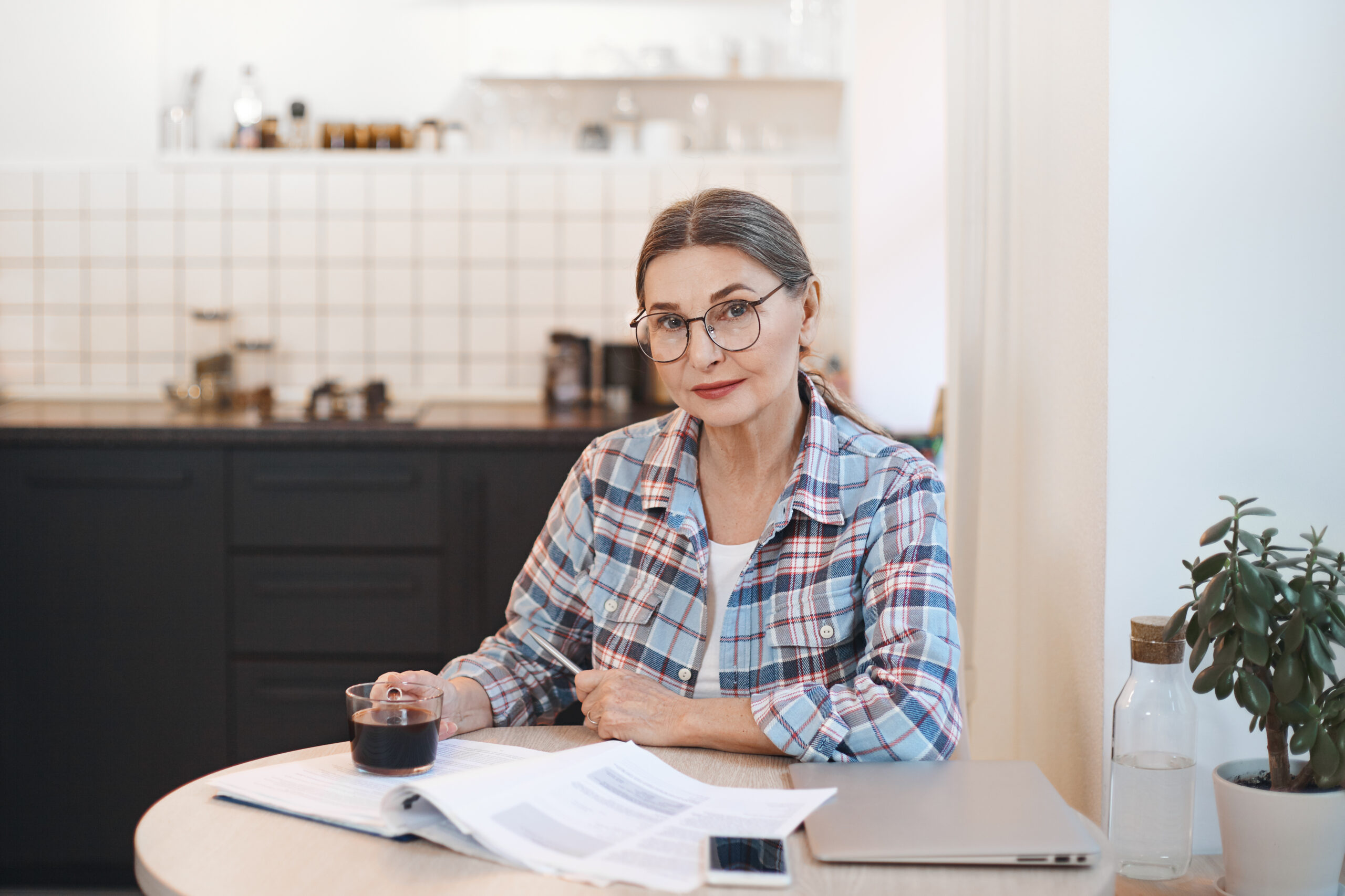 Age, technology, modern lifestyle and occupation concept. Confident female pensioner working remotely from home, sitting in kitchen with laptop computer, notes and cup of coffee, wearing glasses side income after 55